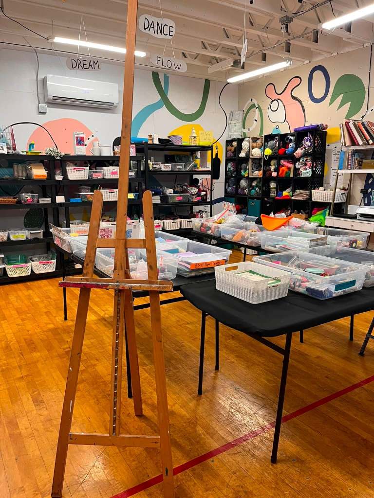 a small art supply store's inventory of shelves with bundles of yarn, bins with art supplies, and book shelves lined with books.